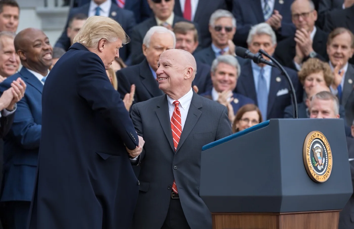 Congressman Brady and President Trump shake hands at a lectern. An audience applauds in the background.
